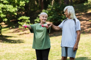 cheerful senior man in sportswear touching hand of wife working out with dumbbells in green park 