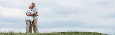 happy senior husband and wife with grey hair hugging and standing on green hill, banner