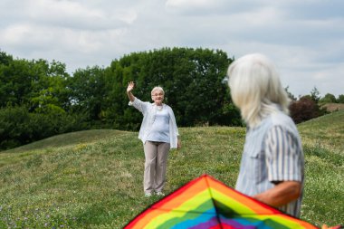 happy senior woman in casual clothes waving hand at husband with kite on green hill 