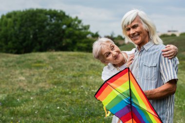 happy senior woman in casual clothes hugging husband with kite on green hill 