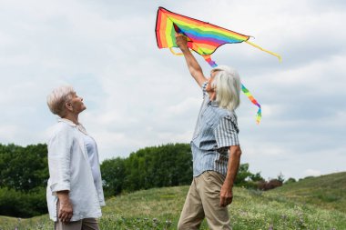 happy senior couple in casual clothes looking at flying kite and standing on green hill 