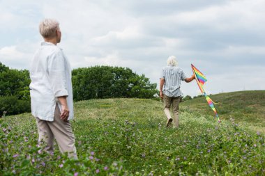 back view of woman with grey hair looking at husband walking with kite on green hill 