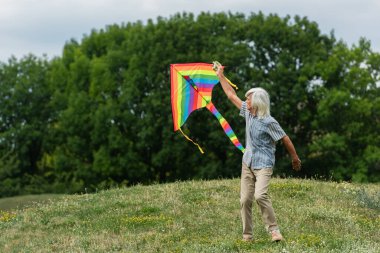 full length of happy senior man in casual clothes playing with kite on green hill 