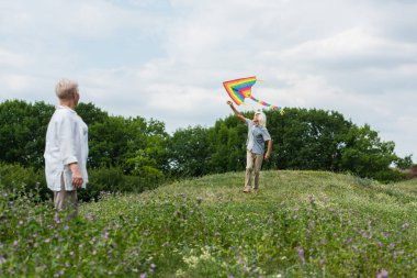 senior woman in casual clothes looking at husband playing with kite on green hill 