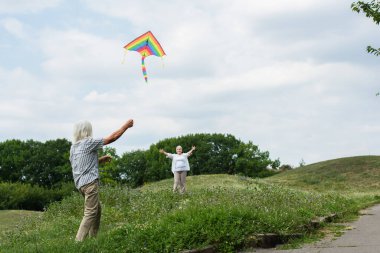 happy senior woman in casual clothes looking at husband playing with kite on green hill 