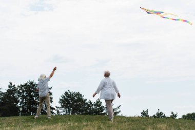 senior woman in casual clothes walking near husband playing with kite on green hill 