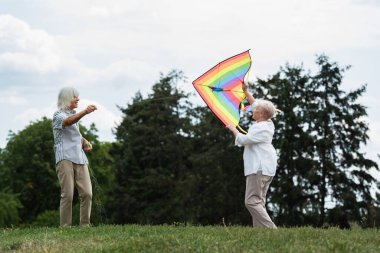 senior woman in casual clothes playing with kite near happy husband on green hill 