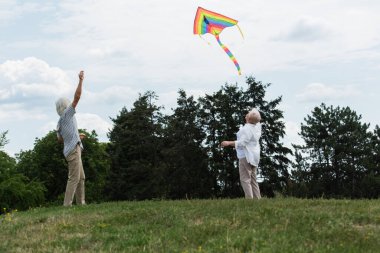 retired couple with grey hair looking at flying kite while standing on green hill 