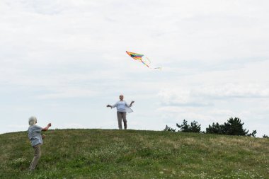 cheerful senior woman in casual clothes looking at kite near husband on green hill 