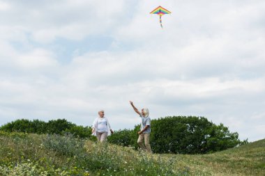 happy senior couple in casual clothes playing with kite on green hill 