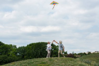 cheerful senior couple in casual clothes holding hands while playing with kite on green hill  clipart