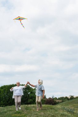 happy senior couple in casual clothes holding hands while playing with kite on green hill 
