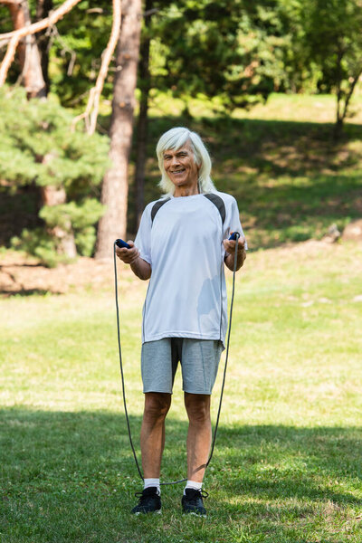 full length of happy senior man with grey hair holding skipping rope in park