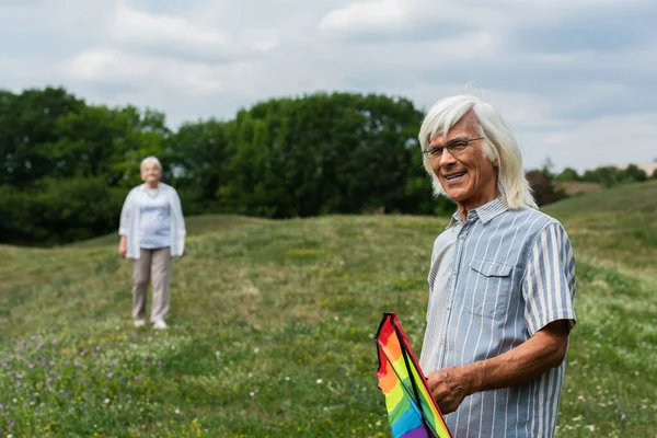 Happy Senior Man Glasses Holding Kite Blurred Wife Standing Green — Fotografia de Stock