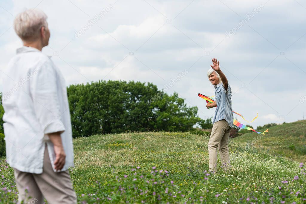 happy senior man in casual clothes holding kite and waving hand at wife while walking on green hill 