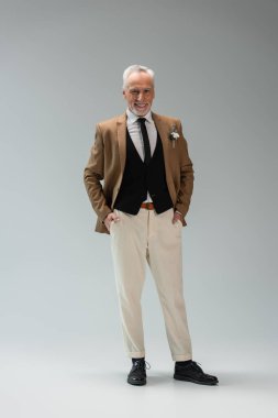 full length of happy middle aged groom in suit standing with hands in pockets on grey 