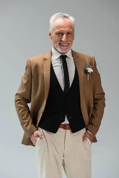happy middle aged groom in suit with floral boutonniere standing with hands in pockets isolated on grey