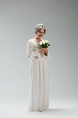 full length of joyful middle aged bride in white wedding dress holding bouquet on grey