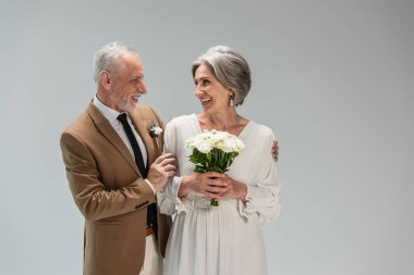 happy mature bride in wedding dress holding bouquet and standing with middle aged groom in suit isolated on grey