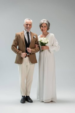 full length of happy mature bride in wedding dress holding bouquet and standing with middle aged groom in suit on grey