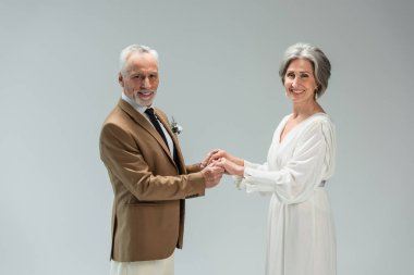 happy mature groom and bride smiling while holding hands isolated on grey 