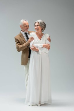 full length of happy middle aged groom hugging cheerful bride in wedding dress on grey