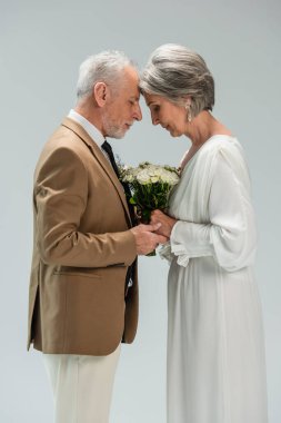 side view of middle aged groom and bride in white dress holding hands near wedding bouquet on grey