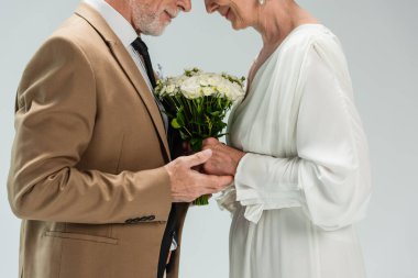 cropped view of middle aged groom and happy bride in white dress holding hands near wedding bouquet on grey