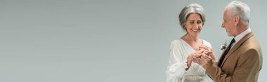 middle aged groom wearing golden engagement ring on happy bride in white dress isolated on grey, banner