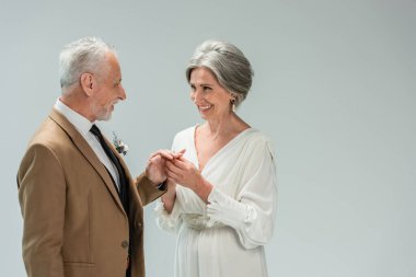happy mature bride in white dress wearing engagement ring on finger of middle aged groom isolated on grey