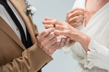 cropped view of groom wearing golden engagement ring on finger of bride in white dress isolated on grey