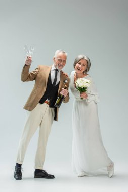 full length of middle aged groom holding bottle of champagne and glasses near happy bride in white dress on grey