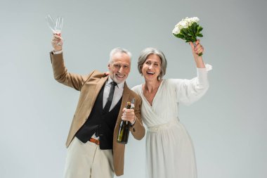 cheerful middle aged groom holding bottle of champagne and glasses near happy bride in white dress isolated on grey