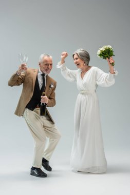 full length of cheerful middle aged man holding bottle of champagne and glasses while dancing with bride in white dress on grey