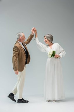 full length of happy middle aged newlyweds dancing while holding hands on grey 
