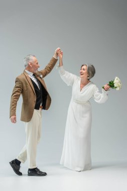 full length of cheerful middle aged newlyweds dancing while holding hands on grey 