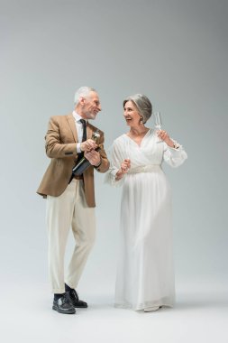 full length of happy middle aged groom opening bottle of champagne near bride in white dress holding glasses on grey 
