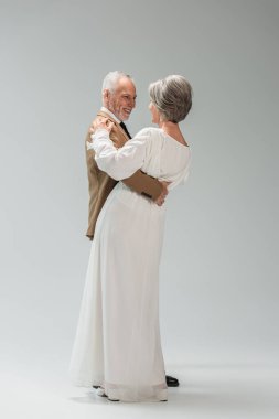 full length of happy mature groom and bride in wedding dress dancing on grey 