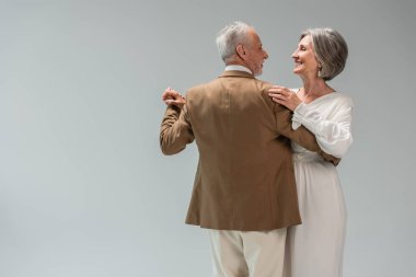 happy middle aged groom and bride in wedding dress holding hands while dancing isolated on grey 