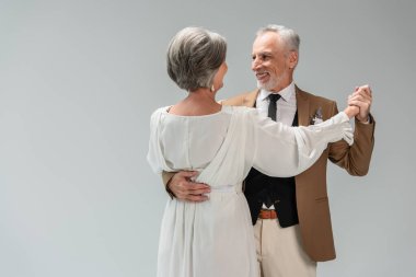 smiling middle aged groom and bride in wedding dress holding hands while dancing isolated on grey 
