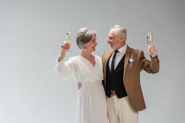 cheerful middle aged groom and happy bride in white dress holding glasses of champagne isolated on grey