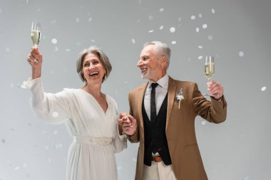 middle aged groom and happy bride in white dress holding glasses of champagne near falling confetti on grey