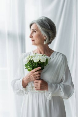 middle aged bride in white dress holding wedding bouquet and standing near curtains and window 