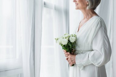 cropped view of happy middle aged bride in white dress holding wedding bouquet and standing near curtains 