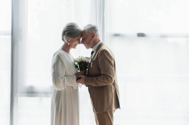 side view of bearded middle aged groom holding wedding bouquet with happy bride in white dress