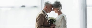 side view of bearded middle aged groom holding wedding bouquet with happy bride in white dress, banner
