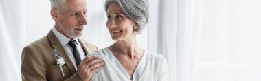 bearded middle aged groom in suit touching shoulder of happy bride in white dress, banner