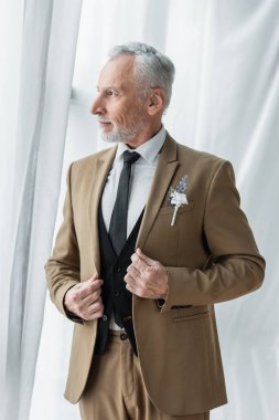 bearded middle aged man in suit with boutonniere posing near white curtains 