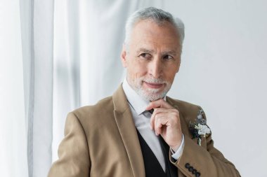 bearded middle aged man in suit with floral boutonniere smiling while looking away near white curtains 
