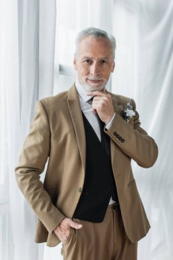 bearded middle aged man in suit with floral boutonniere smiling while posing near white curtains 